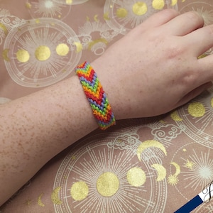 May include: A colourful friendship bracelet with a chevron pattern in red, orange, yellow, green, blue and purple.