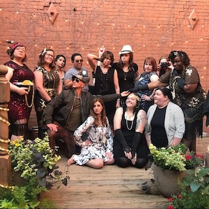 May include: A group of people dressed in vintage attire, posing in front of a brick wall. The individuals are wearing flapper dresses, suits, and accessories like hats and sunglasses. The scene is lit with string lights, suggesting an outdoor event.