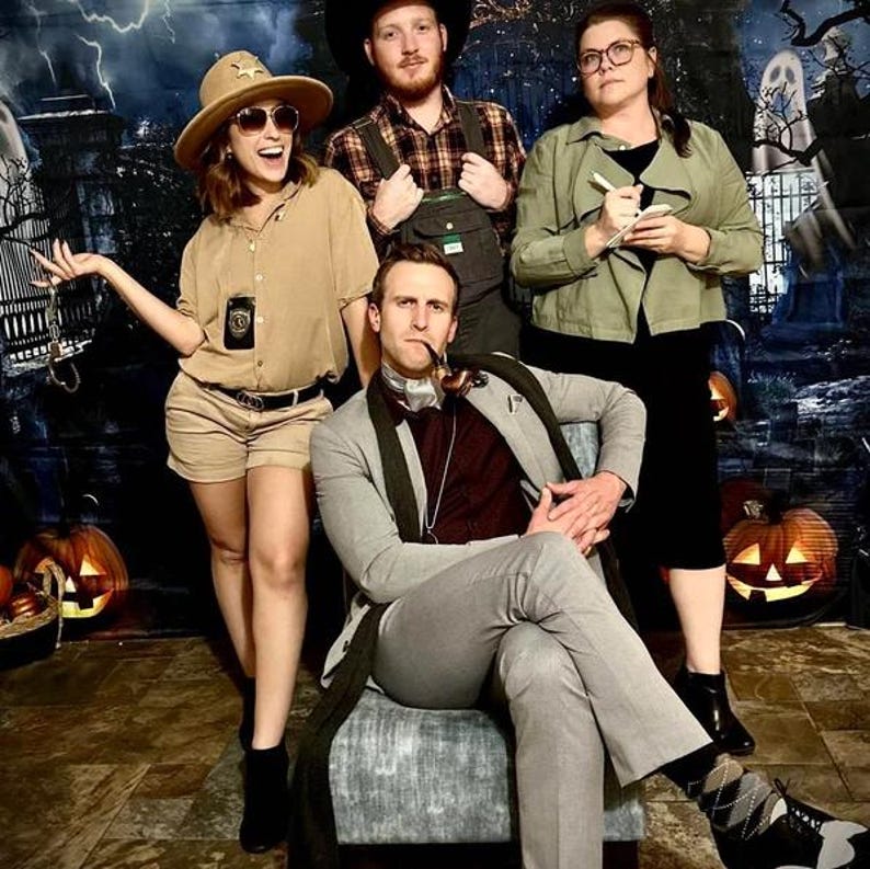 May include: Four people dressed in costumes for a Halloween party. A woman in a brown uniform and a hat with a badge, a man in a plaid shirt and overalls, a woman in a green jacket and a man in a gray suit with a pipe.