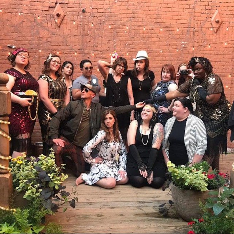 May include: A group of people dressed in vintage attire, posing in front of a brick wall. The individuals are wearing flapper dresses, suits, and accessories like hats and headbands. The scene is lit with string lights, suggesting an outdoor event.