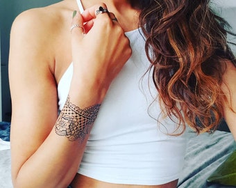May include: A detailed black henna-style temporary tattoo adorns the wrist. The design features intricate floral and geometric patterns. The person is wearing a white halter top and several silver rings. The hair is long and wavy with brown and auburn highlights.