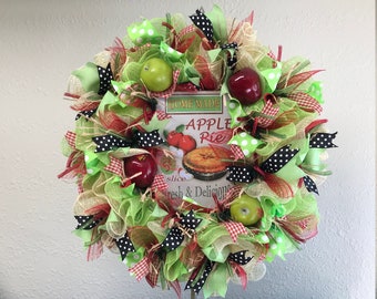 May include: A green, red and black wreath with a sign that says "Home Made Apple Pie 5¢ a slice Fresh & Delicious". The wreath is decorated with green apples, red apples and black and white polka dot ribbon.