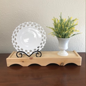 May include: A white ceramic plate with a scalloped edge sits on a wooden shelf with a black metal stand. A white vase with yellow flowers sits on the shelf.