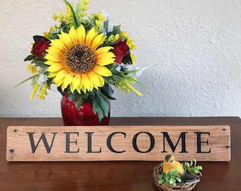 May include: A wooden sign with the word "WELCOME" painted in black letters. The sign is sitting on a wooden table with a red vase filled with yellow sunflowers, red roses, and green foliage. A small yellow bird figurine is sitting in a nest on the table.