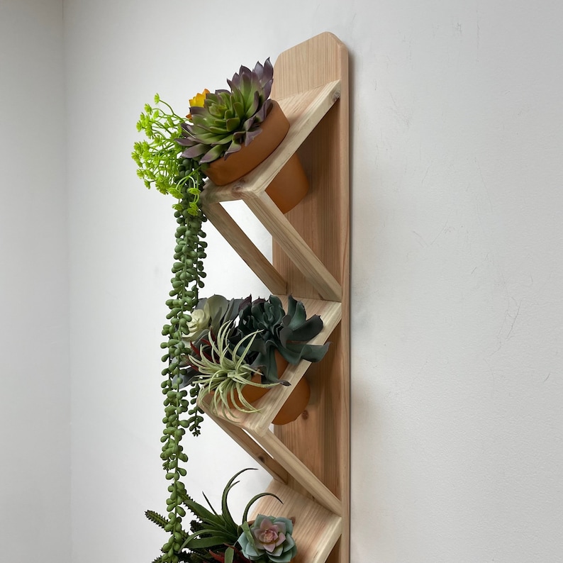 May include: A wooden wall-mounted planter with three shelves, each holding a potted succulent plant. The shelves are angled and create a staggered effect. The planter is made of natural wood and has a light brown finish.