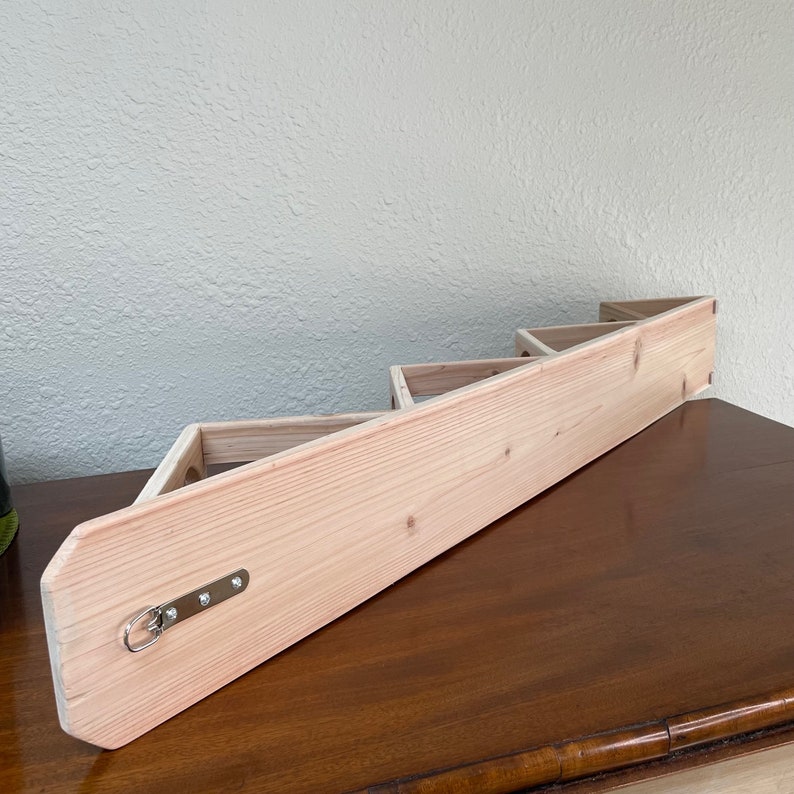 May include: A light wood wall shelf with a triangular design. The shelf has three tiers and is mounted to a wall. The shelf has a metal hook on the bottom.