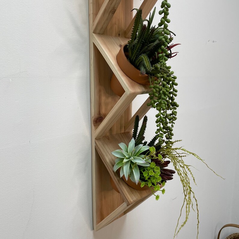 May include: A wooden wall-mounted planter with three triangular shelves. The shelves are filled with potted succulents and plants. The planter is made of natural wood and has a rustic look.