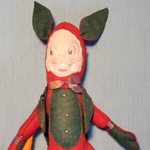 Può includere: Un coniglio di peluche vintage in feltro che indossa un vestito rosso e verde con un papillon. La bambola ha un viso dipinto e tiene una carota in mano.
