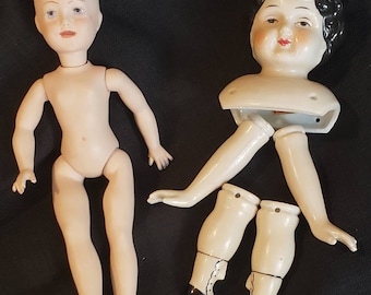 Op de afbeelding: Twee porseleinen poppen, één met een volledig lichaam en de andere met een hoofd, armen en benen. De pop met een volledig lichaam is naakt en heeft zwart haar. De pop met alleen een hoofd, armen en benen heeft zwart haar en draagt zwarte schoenen.
