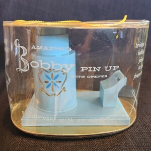 Könnte beinhalten: Ein Vintage "Bobby Pin Up" Spender mit Öffner, in einer transparenten Kunststoffverpackung. Der Spender ist hellblau mit goldenen Akzenten und Text, darunter "Bobby" und "Amazing". Der Sockel ist hellblau und gold.