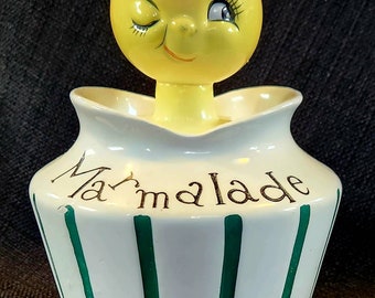 May include: A white ceramic jar with green stripes and the word "Marmalade" written in gold lettering. The jar has a yellow ceramic lemon with a winking face on top.