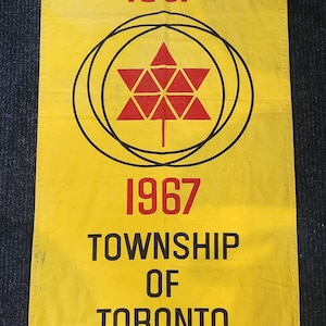 Puede incluir: Una pancarta de tela amarilla con texto negro que dice "Confederation Centennial 1867 1967 Township of Toronto". Una hoja de arce estilizada roja con un contorno negro está centrada en la pancarta. La pancarta tiene un fondo puntiagudo con el texto "Side A" en negro.