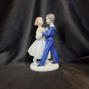 Könnte beinhalten: Porzellanfigur, die ein tanzendes Paar darstellt. Die männliche Figur trägt einen blauen Anzug, die weibliche ein weißes Kleid. Der Sockel ist weiß und oval. Die Figur ist ein dekorativer Artikel.
