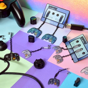 May include: A set of dangle earrings featuring miniature GameCube controllers. The earrings are made of black metal with a silver chain and a variety of colored controllers. The earrings are displayed on a pink, blue, and purple background. The text "GameCube Earrings" is visible on the image.