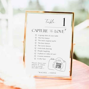 以下が含まれることがあります： 「Table 1」と「Capture the Love」の文字が入った金色のフレームのテーブルサイン。サインには写真のプロンプトのリストと、写真やビデオを共有するためのQRコードが含まれています。サインは木製の表面にあります。