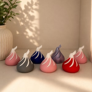 May include: A collection of colorful, decorative objects shaped like a Hershey's Kiss. The objects are in various colors, including pink, gray, blue, purple, and red, with white accents. They are arranged on a white shelf.