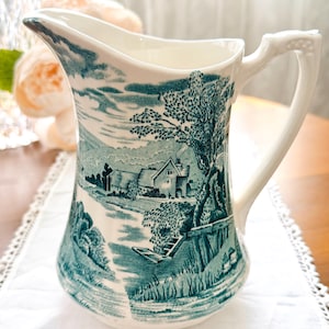 Peut inclure: Un pichet en céramique couleur crème orné d'un motif bleu sarcelle représentant un paysage rural avec un cottage, des arbres et une rivière. Le pichet présente une anse incurvée, un large bec verseur et une finition lisse et brillante.
