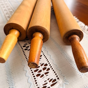 May include: Three cylindrical wooden rolling pins with light brown barrels and darker brown handles. Each rolling pin features handwritten black text on the end of the barrel, showing distinct wood grain patterns and smooth, polished surfaces.