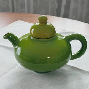 May include: A glossy, grass-green ceramic teapot with a rounded body, a curved handle, a short spout, and a matching lid featuring a small, round knob handle on top. The teapot has a smooth, reflective surface and a uniform, vibrant green colour.