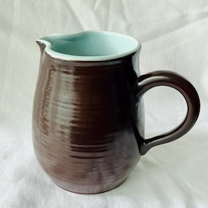 May include: A ceramic pitcher with a glossy dark brown exterior and a smooth light blue interior. The vessel features a rounded body, a curved handle on the right side, and a small pouring spout at the top rim, showcasing a handcrafted finish.
