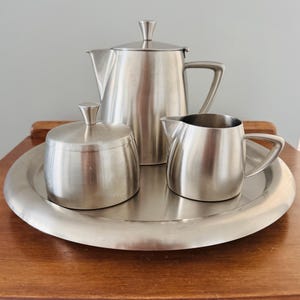May include: A silver-toned stainless steel tea service set consisting of a teapot with a lid, a lidded sugar bowl, and a creamer jug, all arranged on a matching round serving tray. Each piece features a smooth, brushed metallic finish.