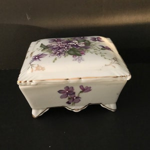 May include: A square white porcelain trinket box with a hinged lid. The lid is decorated with a floral design featuring purple and green flowers. The box has gold trim around the edges and sits on four small feet.