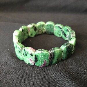 May include: A bracelet made of green and black rectangular stones with red accents. The stones are arranged in a circular pattern, and the bracelet is set against a black background.