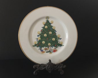 May include: A white ceramic Christmas plate with a gold rim features a green Christmas tree with red and blue ornaments, white lights, and a star on top.  There are small figures of children and toys at the base of the tree.