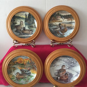 May include: Set of four decorative plates featuring a wood frame and a painted image of ducks in various settings. The plates are displayed on small gold metal stands.
