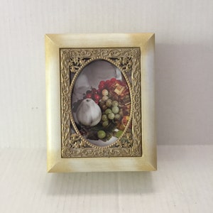 May include: A gold-toned picture frame with an ornate design. The frame contains a picture of a still life with fruit, including a pear, grapes, and apples.