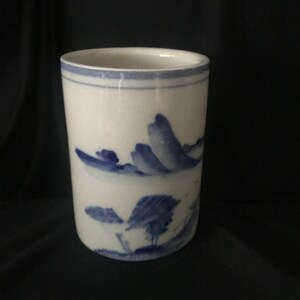 May include: White ceramic tumbler with a blue landscape design. The design features stylized mountains, trees, and water. The rim of the tumbler has a thin blue band. The tumbler is cylindrical in shape.
