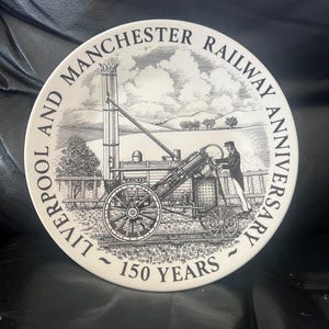 May include: A white decorative plate with a black and white illustration of a steam engine and the text "LIVERPOOL AND MANCHESTER RAILWAY ANNIVERSARY ~ 150 YEARS". The plate has a black border.