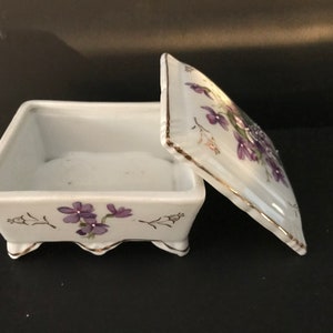 May include: A white porcelain trinket box with a hinged lid. The box is decorated with purple flowers and gold trim. The box has four small feet.