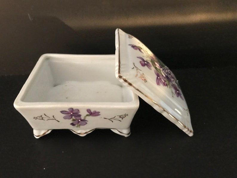 May include: A white porcelain trinket box with a hinged lid. The box is decorated with purple flowers and gold trim. The box has four small feet.