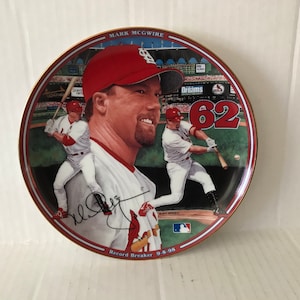 May include: A commemorative plate featuring a baseball player in a red uniform with the number 62 on it. The plate has a gold rim and the text "Mark McGwire Record Breaker 0-9-98" on the bottom.