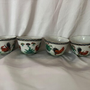 May include: Four white ceramic bowls with a hand-painted rooster design. Each bowl features a red rooster, green foliage, and a black and gold rim. The bowls are set against a white background.