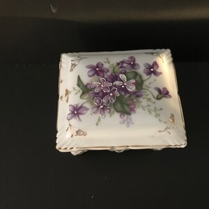 May include: A square white porcelain trinket box with a floral design. The box features a delicate floral pattern with purple violets and green leaves, accented with gold trim.