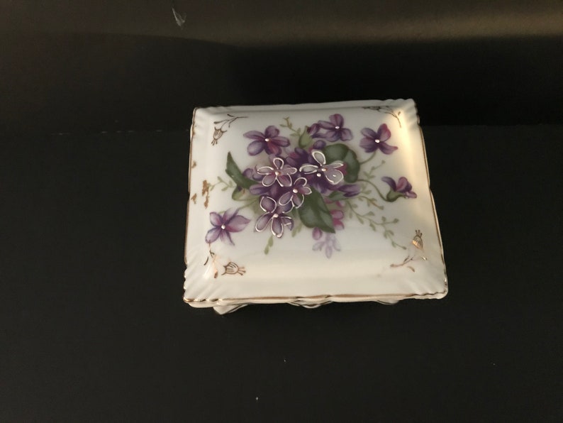 May include: A square white porcelain trinket box with a floral design. The box features a delicate floral pattern with purple violets and green leaves, accented with gold trim.