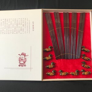 Pode incluir: Um conjunto de pauzinhos de madeira numa caixa vermelha com forro de veludo vermelho. Os pauzinhos são decorados com letras douradas e a caixa tem uma pequena ilustração de uma pessoa. A caixa também tem texto chinês na frente.