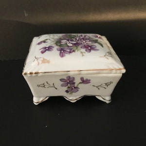 May include: A white porcelain trinket box with a square shape and a floral design. The lid features a cluster of purple flowers with green leaves, while the base has a smaller floral design. The box has gold trim around the edges and four small feet.
