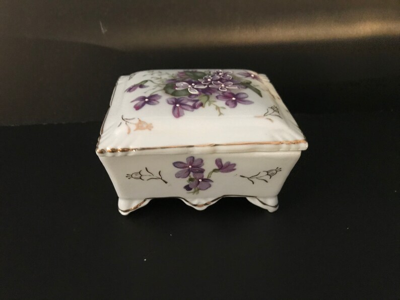 May include: A white porcelain trinket box with a square shape and a floral design. The lid features a cluster of purple flowers with green leaves, while the base has a smaller floral design. The box has gold trim around the edges and four small feet.