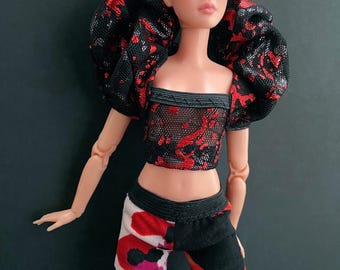 May include: A fashion doll with short dark hair and a stylish outfit. The doll wears a black crop top with red splatter design and matching puffy sleeves. The doll's pants are split, with a red and white pattern on one side and black on the other.