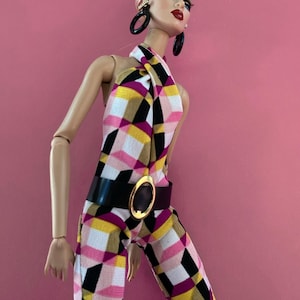 May include: A fashion doll with light pink hair, wearing large black hoop earrings and a halter-neck jumpsuit featuring a geometric pattern in pink, yellow, black, and white. The jumpsuit is cinched at the waist with a black belt featuring a gold oval buckle.