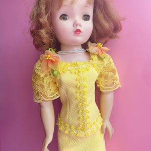 May include: A vintage doll with auburn hair and large eyes, wearing a yellow dress with lace sleeves and floral accents. The dress is adorned with white beaded trim and a matching skirt. A pearl necklace completes the ensemble. The doll is posed against a pink background.