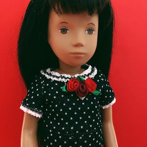 May include: A doll with black hair and brown eyes wearing a black dress with white polka dots. The dress has white lace trim and is adorned with red fabric roses and green leaves. The doll is set against a red background.