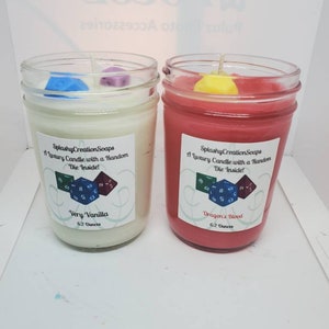 May include: Two glass jars with lids containing candles. The candles are white and red. Each jar has a label that says "SplashyCreationSoaps" and "A Luxury Candle with a Random Die Inside!" The white candle jar has a label that says "Very Vanilla" and the red candle jar has a label that says "Dragon's Blood". Both jars are 6.2 ounces.