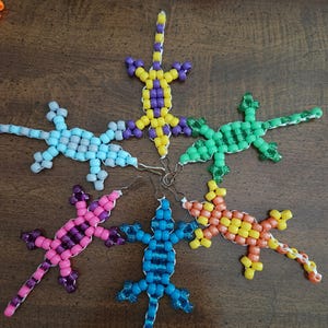 Könnte beinhalten: Sechs echsenförmige Ornamente aus bunten Kunststoffperlen, die in einem Kreis angeordnet sind. Jede Echse weist eine einzigartige Farbkombination auf, darunter Gelb und Lila, Hellblau, Grün, Pink, Dunkelblau und Orange, alle mit Metallhaken versehen.