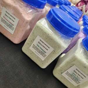 Puede incluir: Tres frascos de plástico transparente con tapas azules llenos de sales de baño. Los frascos están etiquetados con el texto "Splashy CreationSoaps Luxury Bath Soak" y los nombres de las fragancias: "Moroccan Cashmere", "Wild Wilderness" y "Green Tea & Lemongrass".