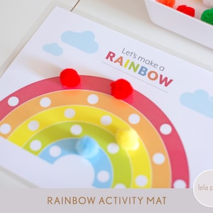 May include: A rainbow-colored activity mat with white circles for pom-poms. The mat has the text "Let's make a RAINBOW" at the top. There are pom-poms in red, yellow, and blue on the mat.