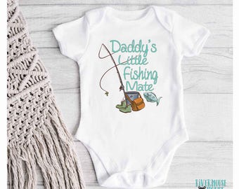 Könnte beinhalten: Weißer Baby-Strampler mit einer türkisfarbenen Grafik einer Angelrute, einem Eimer und einem Fisch. Der Text "Daddy's Little Fishing Mate" ist in Türkis geschrieben.